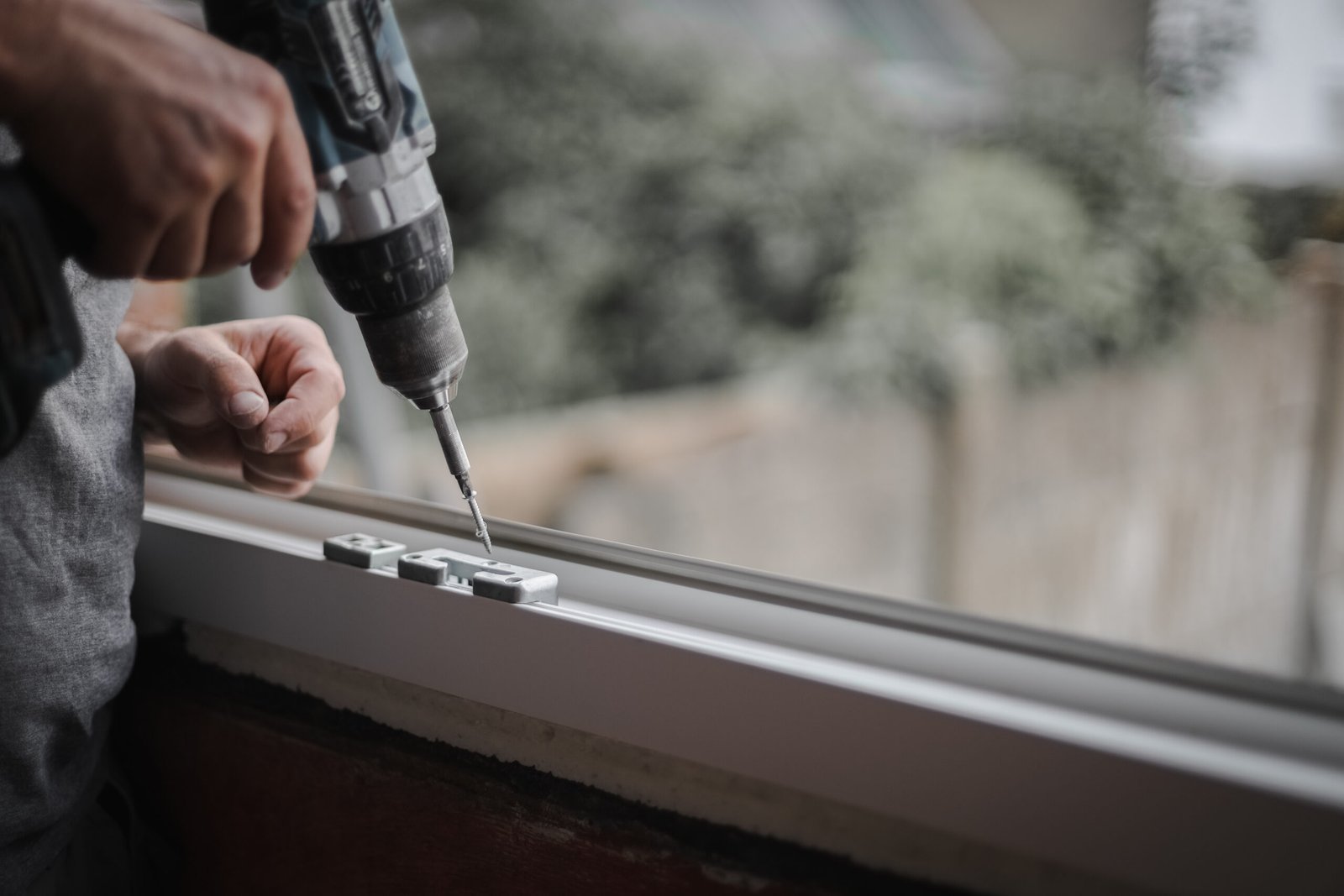 Young caucasian man holding a drill with a screw over the fittings of a plastic window frame in a room where renovations are taking place with copy space on the right, close-up side view with selective focus. The concept of home renovation, washing window frames.
